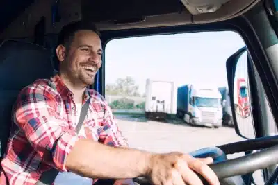 Hombre sonriente conduciendo un camión, visto desde el interior de la cabina. Lleva puesta una camisa de cuadros rojos y el cinturón de seguridad. Al fondo se pueden ver otros camiones estacionados en un área de descanso.