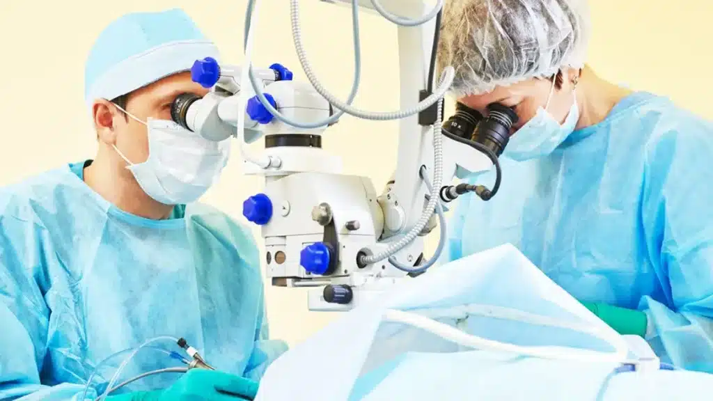 Dos profesionales sanitarios con bata y mascarilla realizan una operación con microscopio quirúrgico en un quirófano.