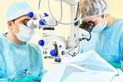 Dos profesionales sanitarios con bata y mascarilla realizan una operación con microscopio quirúrgico en un quirófano.