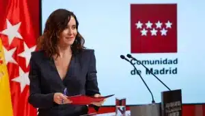 Isabel Díaz Ayuso durante una comparecencia oficial de la Comunidad de Madrid, con el logo institucional y las banderas de España y Madrid de fondo.