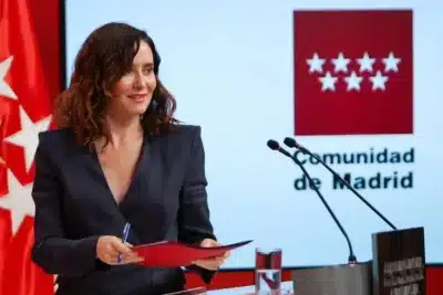 Isabel Díaz Ayuso durante una comparecencia oficial de la Comunidad de Madrid, con el logo institucional y las banderas de España y Madrid de fondo.